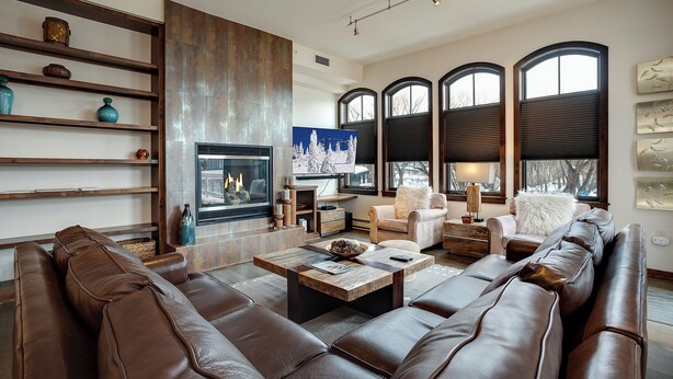 A modern living room with a fireplace, leather sofas, wooden shelves, and large windows.