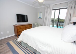 Ein Schlafzimmer mit einem großen Bett, einem an der Wand befestigten Fernseher, einer Holzkommode und Blick auf einen Pool durch ein Fenster.