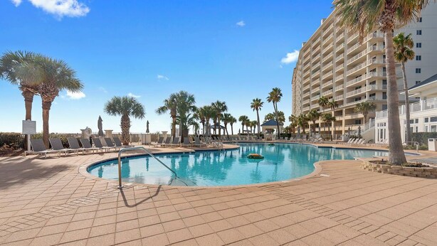 A pool area with lounge chairs and palm trees, surrounded by a high-rise building. - Saltwater Cottage, A Cozy Beach Club Cottage-Cottage 21- Signature Properties (Gulf Shores)