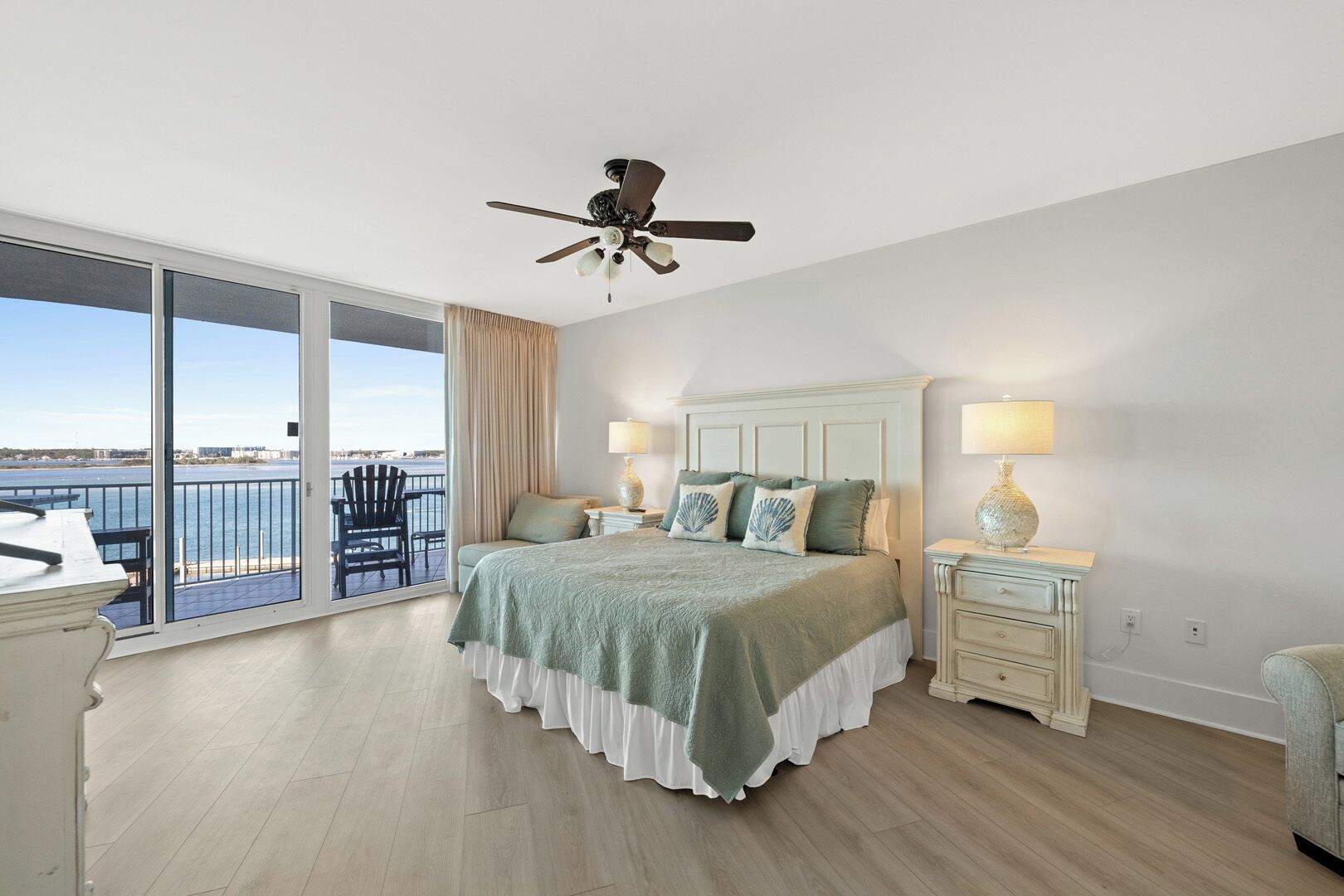 A bedroom with a large bed, two bedside tables, a ceiling fan, and a view of the sea through a sliding glass door.