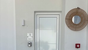 A white door with a frosted glass panel, a wall-mounted intercom, a red fire alarm, a woven mirror, and a potted plant. - Agathi Beauty Studios (Rhodes)