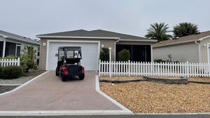 A residential area with a driveway leading to a garage, a white picket fence, and a row of similar houses. - Stylish Home in Duval w/Granite, Tile Floors, Glass Lanai & Gas Golf Cart! (The Villages)