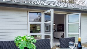 An open sliding glass door leading to a patio area with a table set for two, a potted plant, and a wine bottle. - 4 Star Holiday Home in Borkop (Borkop)