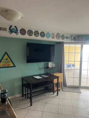 A room with a television mounted on the wall, a table with a laptop, and a beach-themed wall decoration.