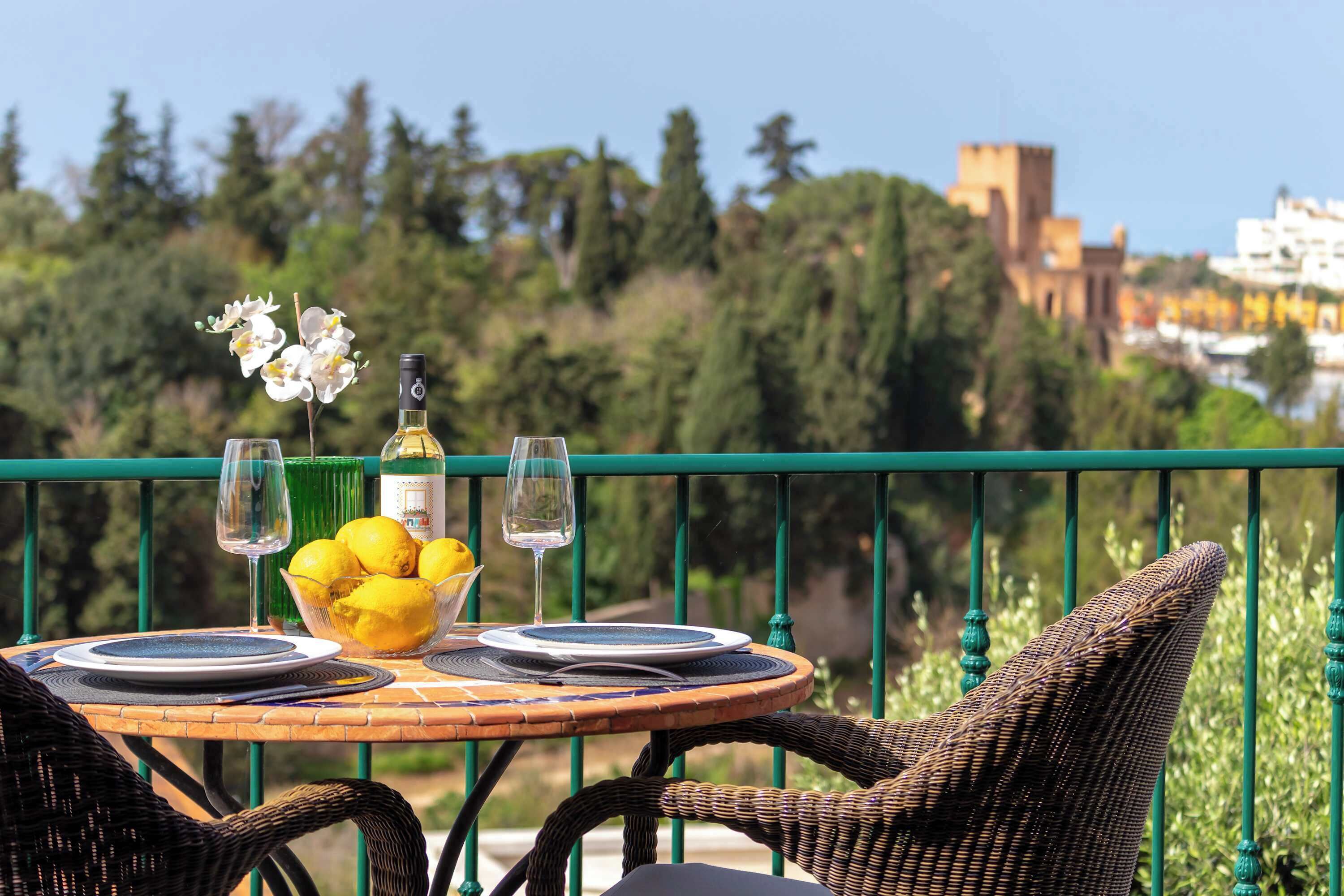 A table set with plates, glasses, a bottle, and lemons on a balcony with a view of greenery and buildings.