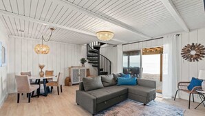 Una sala de estar moderna con un sofá seccional gris, una mesa de comedor con sillas, una araña y una escalera que conduce a un balcón con vistas.