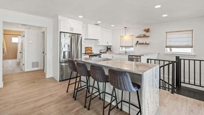 Una cocina moderna con una isla central, taburetes de bar, electrodomésticos de acero inoxidable y vistas al salón.