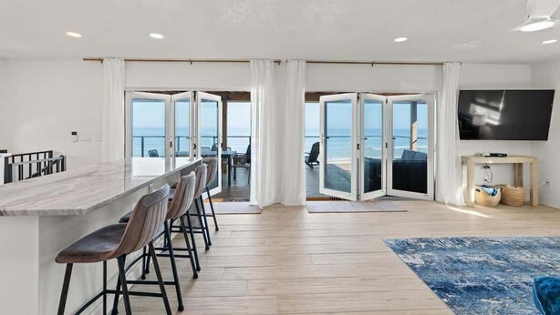 Un salón moderno con televisor grande, una zona de bar con taburetes y vistas al océano a través de puertas abiertas.