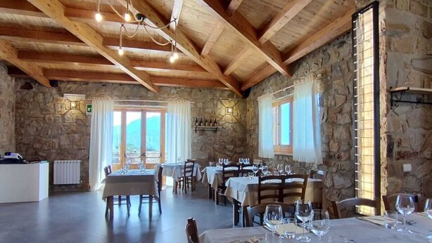 A restaurant with wooden beams and stone walls, tables set with wine glasses and cutlery, and a view of the outdoors. - Foresteria Mongerrati (Isnello)