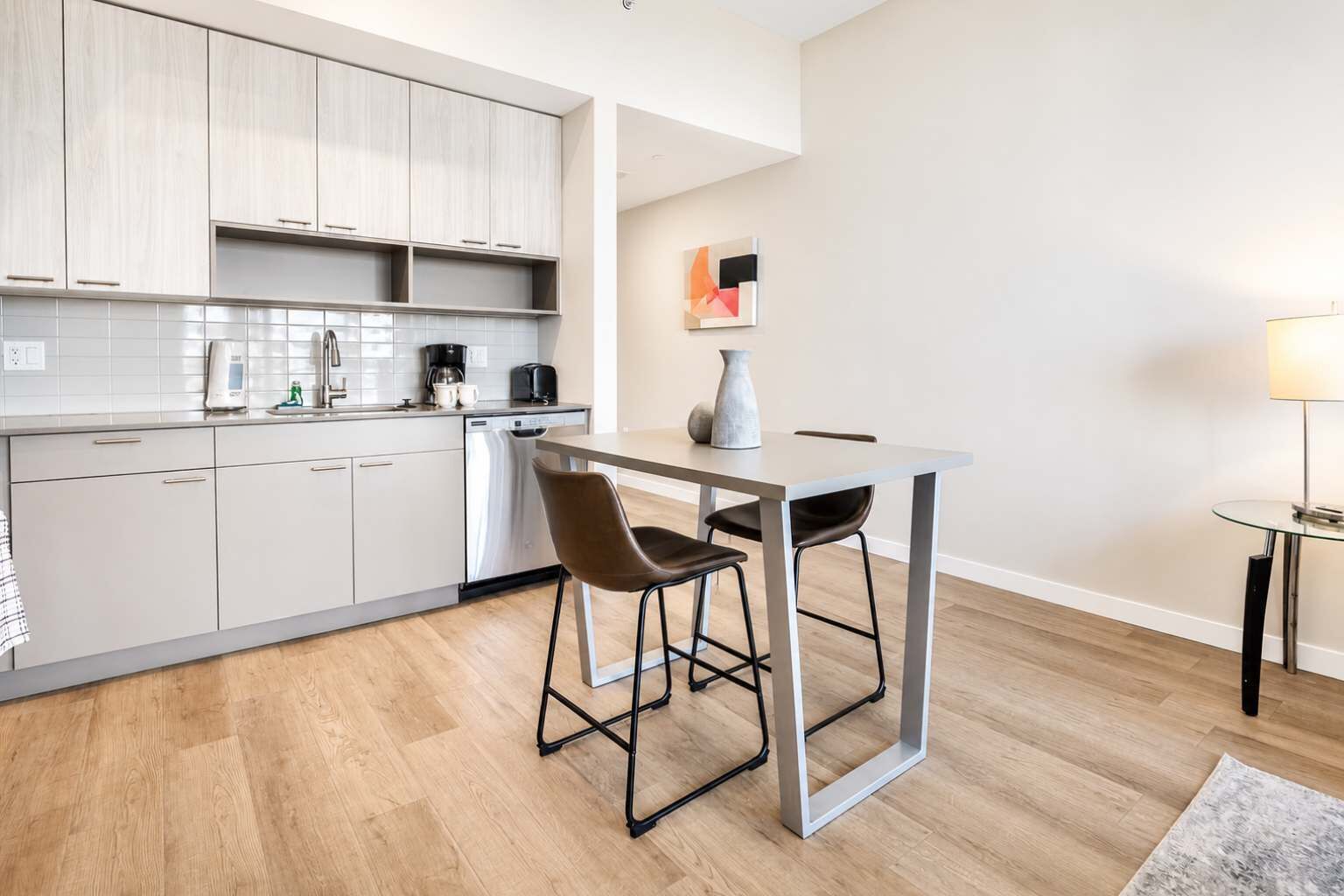 A modern kitchen with white cabinets, a dishwasher, a coffee maker, and a dining area with a table and two chairs.