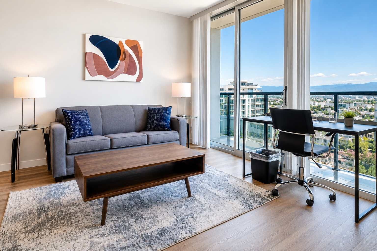A modern living room with a sofa, coffee table, and a desk with a chair. There is a large window with a view of buildings and a mountain range.