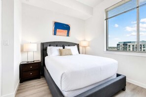 Une chambre d’hôtel moderne avec un grand lit, des lampes de chevet, une vue sur la ville et une œuvre d’art encadrée au mur.