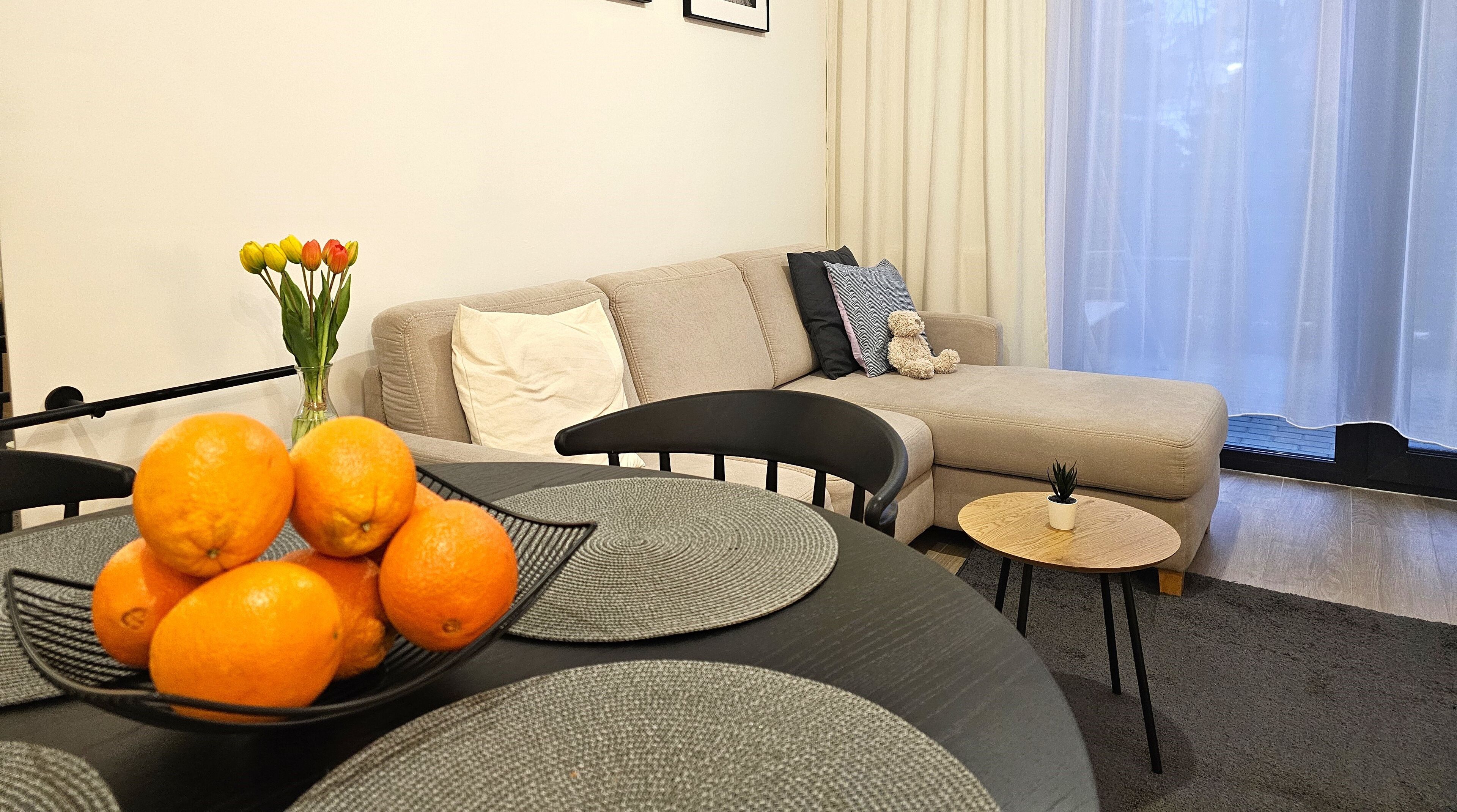 Ein modernes Wohnzimmer mit einem grauen Sofa, einem kleinen runden Tisch und einem Esstisch mit einer Schüssel Orangen.
