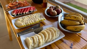 A breakfast buffet with various fruits and pastries on a wooden table by a window. - Hospedaria Paiol (São Bento do Sapucaí)