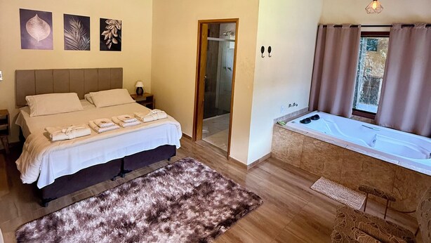 A bedroom with a large bed, a wooden ceiling, a small bathroom, and a window with curtains. - Hospedaria Paiol (São Bento do Sapucaí)