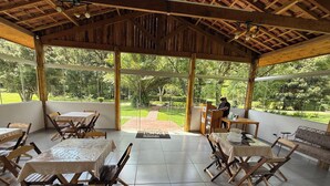 An outdoor dining area with wooden tables and chairs, a person at a podium, and a view of a lush garden. - Hospedaria Paiol (São Bento do Sapucaí)