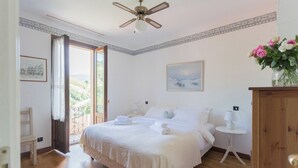 A bedroom with a bed, bedside tables, a ceiling fan, a painting, and a view through an open door. - Vacation Family Home in Tremezzo by Rent all Como (Tremezzina)