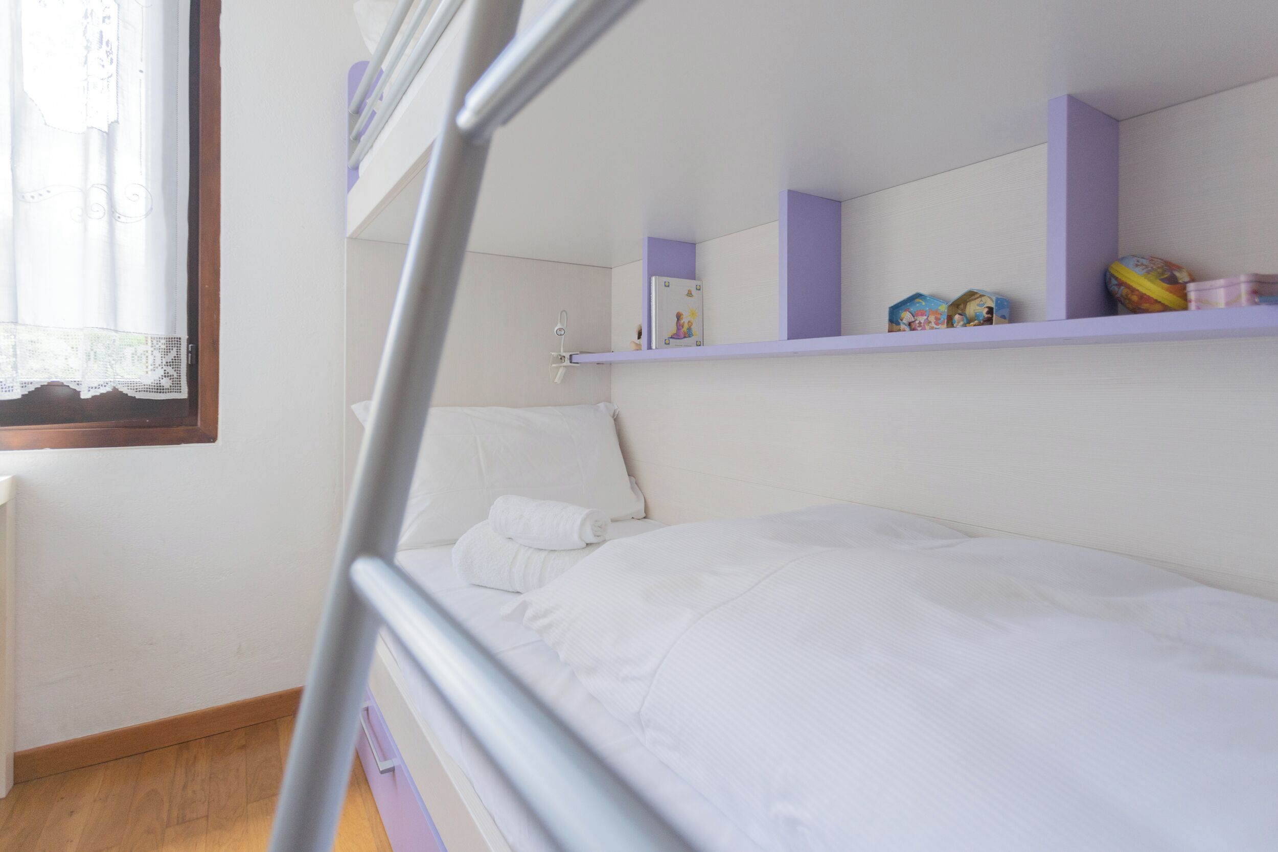 Habitación con literas, una ventana, un estante con juguetes y una cama blanca.