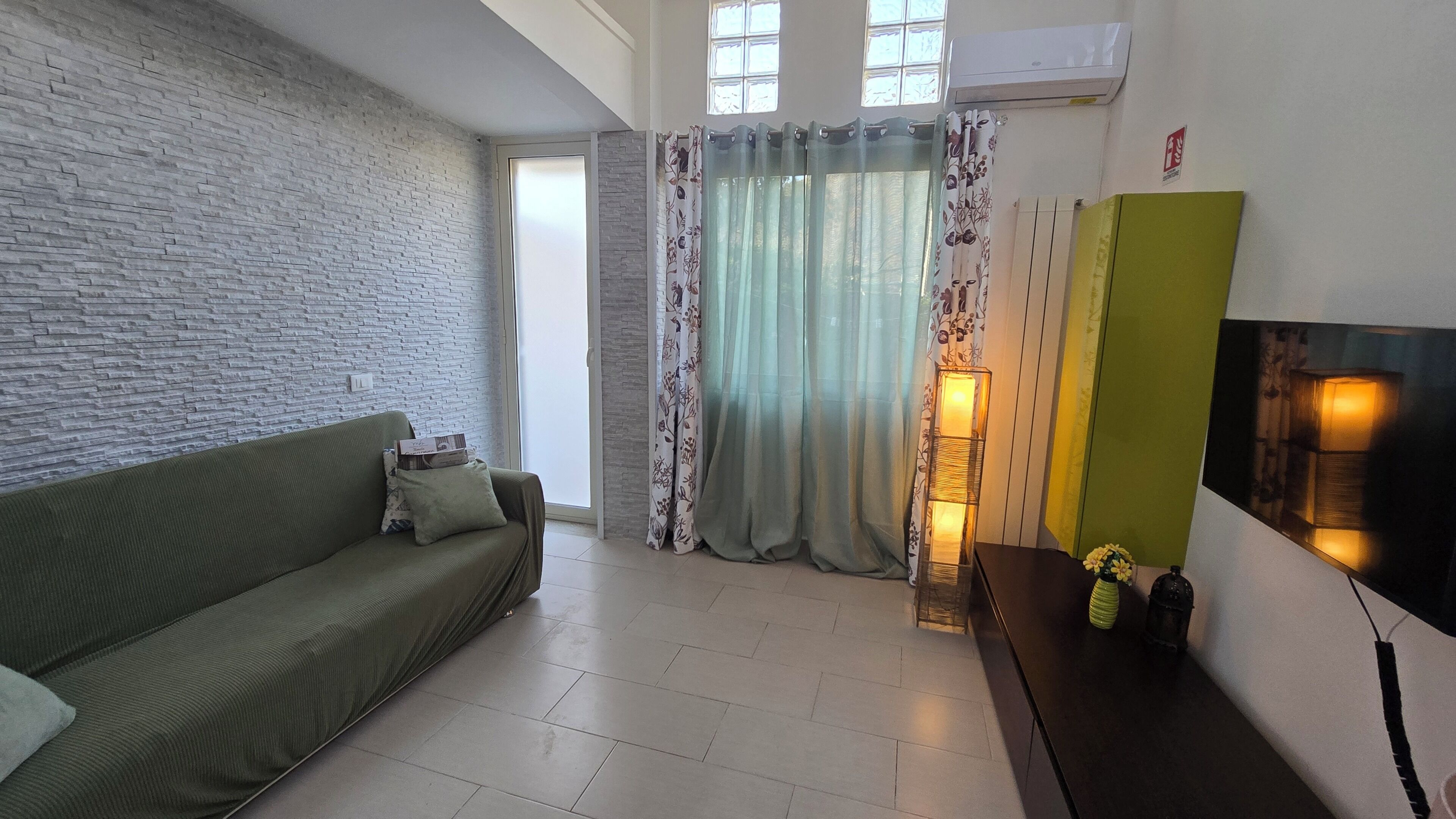 Un salón con un sofá verde, una mesa de centro de madera, un televisor montado en la pared y una ventana con cortinas.