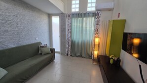 Ein Wohnzimmer mit einem grünen Sofa, einem Holztisch, einem an der Wand befestigten Fernseher und einem Fenster mit Vorhängen.
