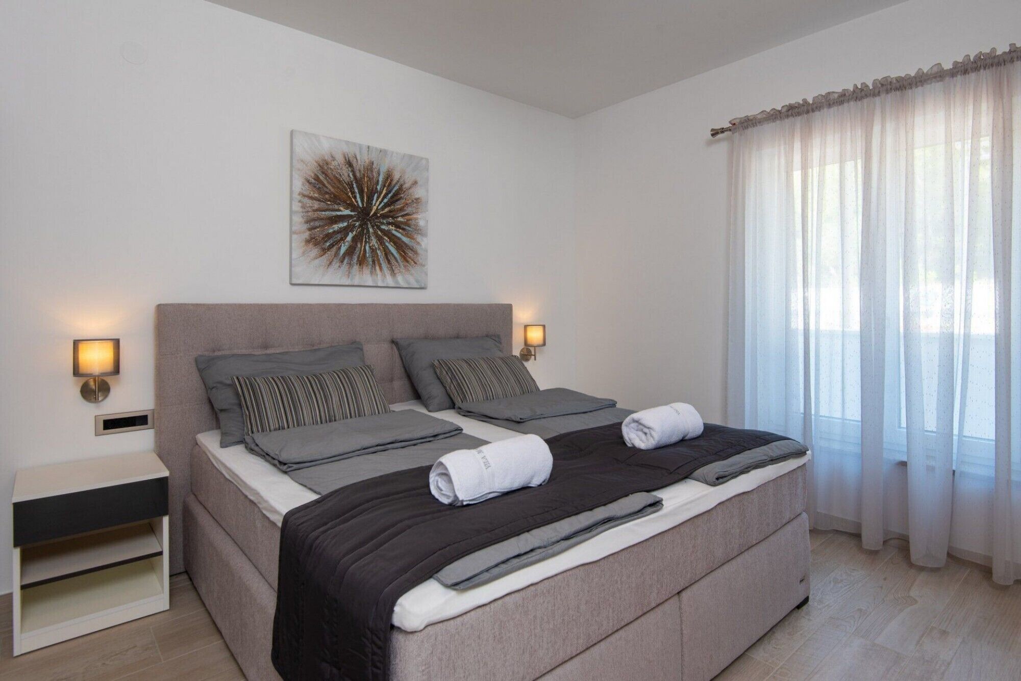Um quarto moderno com uma cama grande, duas luminárias de cabeceira, uma obra de arte na parede e cortinas leves.