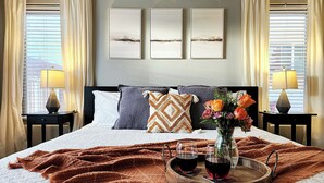 Ein ordentlich bezogenes Bett mit einer braunen Decke, ein Tablett mit Weingläsern und eine Blumenvase, sowie Kunstwerke an der Wand.