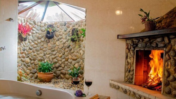 A bathroom with a stone wall, a bathtub, and a stone fireplace with a fire. - Pousada Pau Brasil (Maringá)