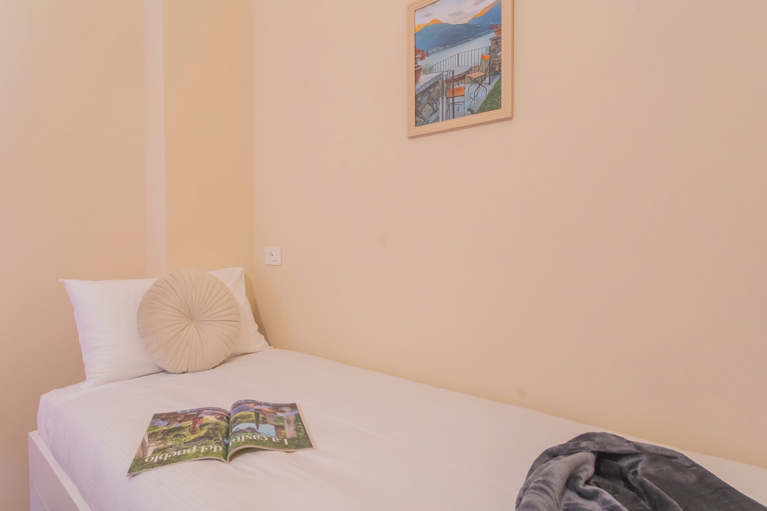 A single bed with a white mattress and two pillows, a framed picture on the wall, and a book on the bed.