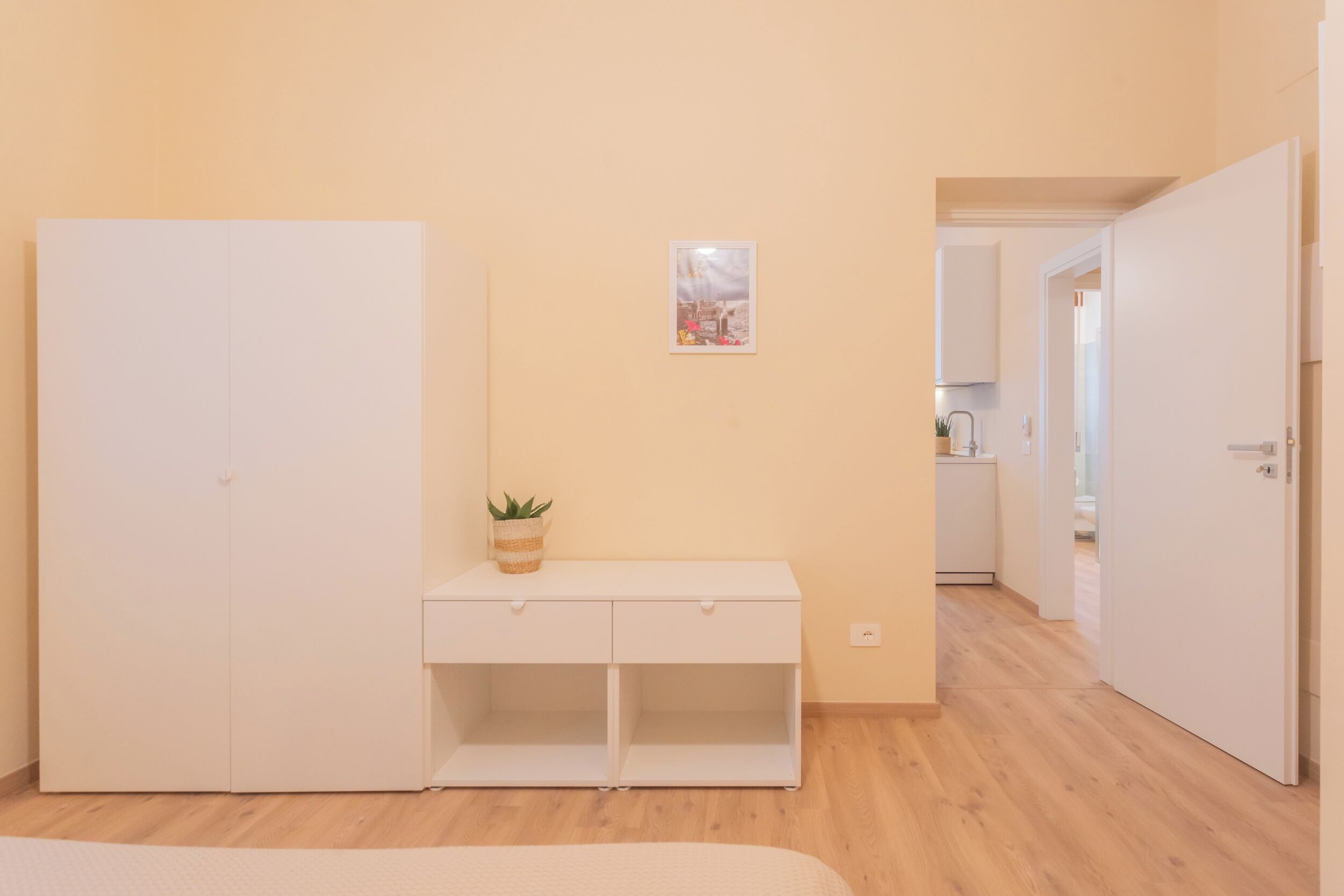 Une chambre à coucher avec un lit, une armoire blanche, une petite table de chevet avec une plante, et une porte donnant sur une salle de bain.