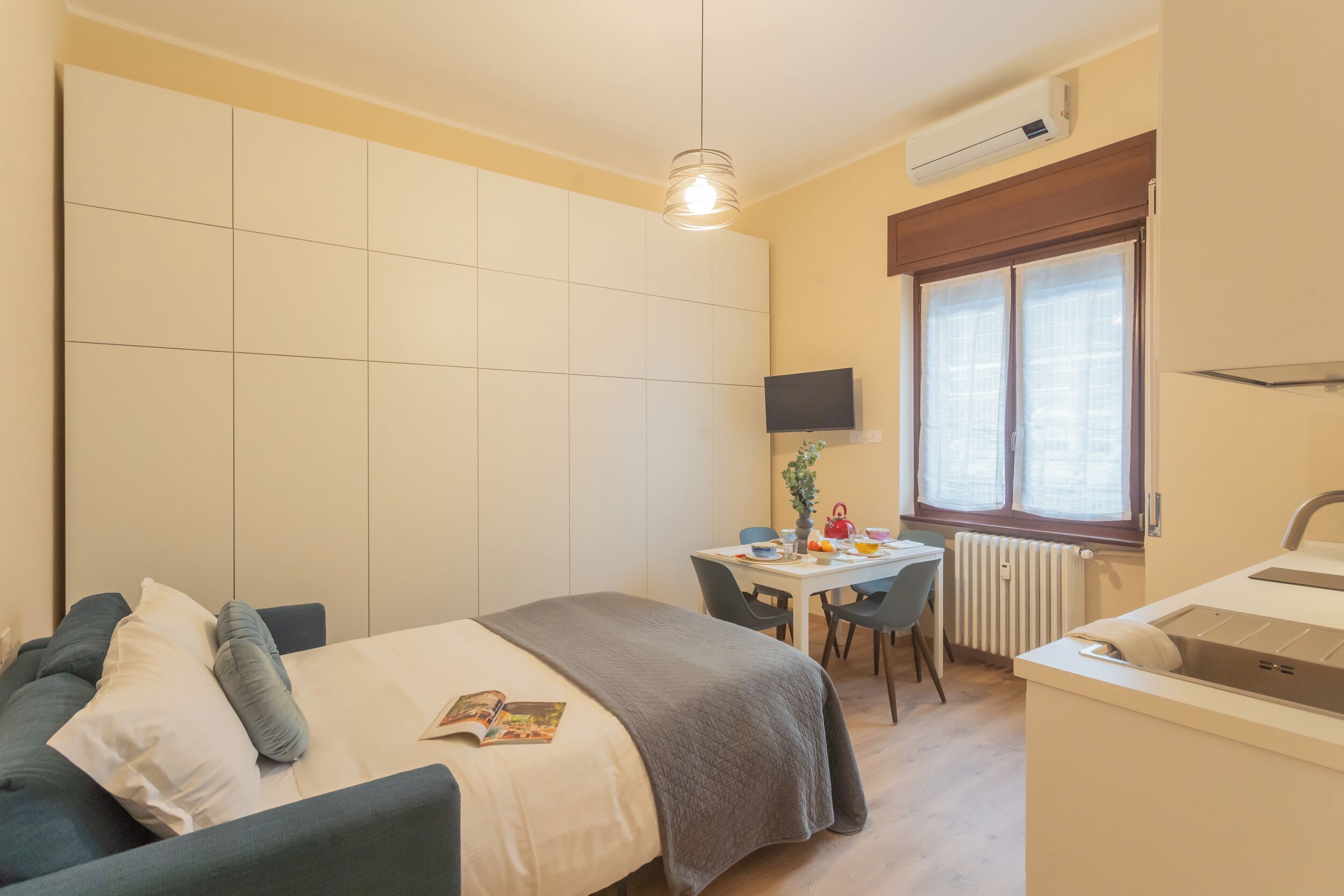 A bedroom with a bed, a desk, a chair, a TV, and a window with blinds.