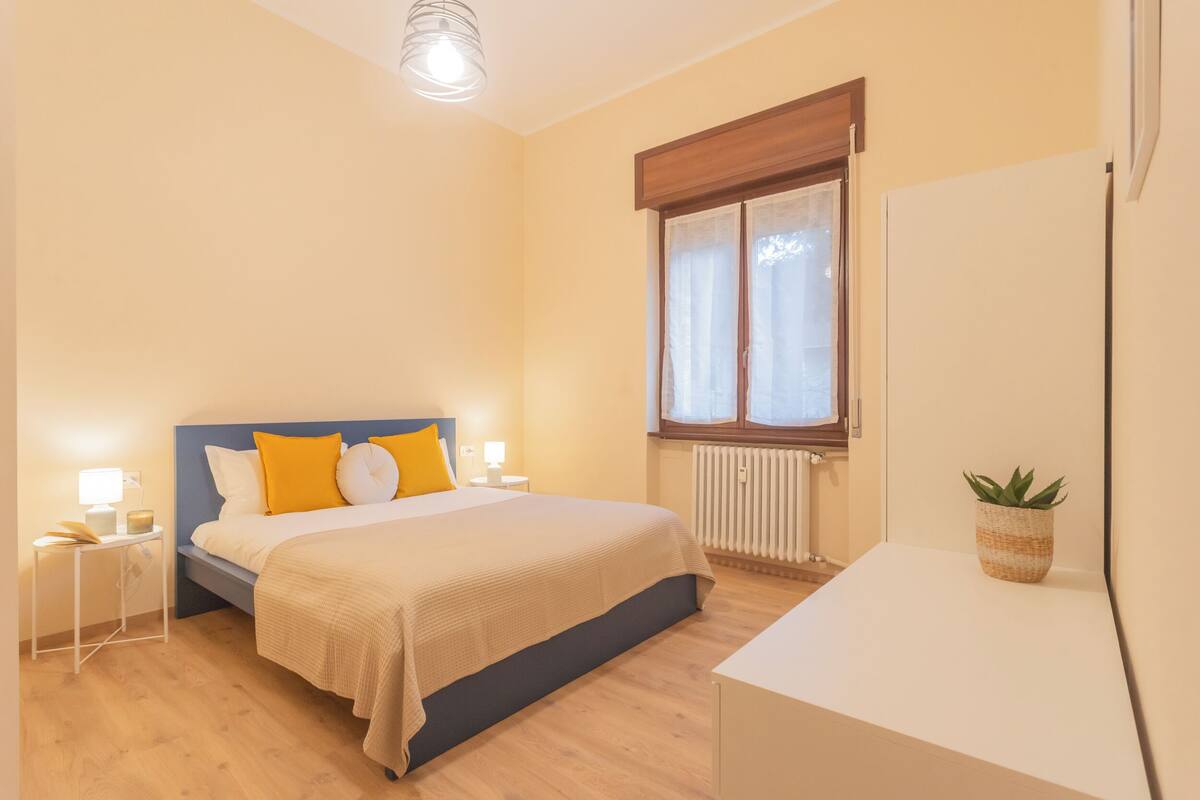 Een slaapkamer met een bed, nachtkastjes, een raam met gordijnen, een radiator en een kleine plant op een tafel.
