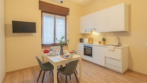 Una cucina compatta con mobili bianchi, un forno a microonde e un lavandino. C'è un tavolo da pranzo con sedie e una finestra con tende.