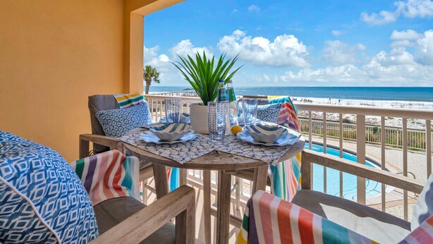 Uma varanda com vista para a praia, uma mesa com almofadas estampadas e um vaso de planta.