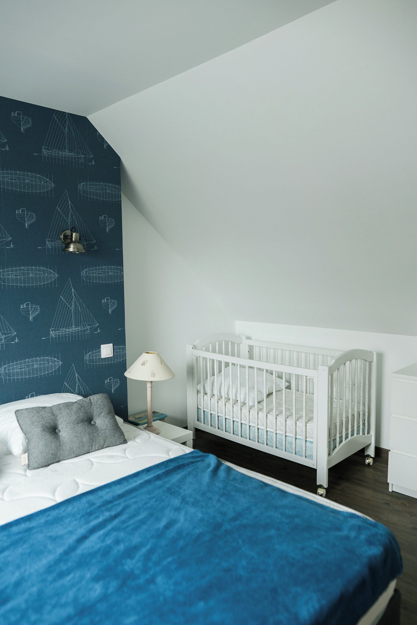 A bedroom with a white crib, a bed with a blue blanket, a nightstand with a lamp, and a wall with a nautical-themed wallpaper.