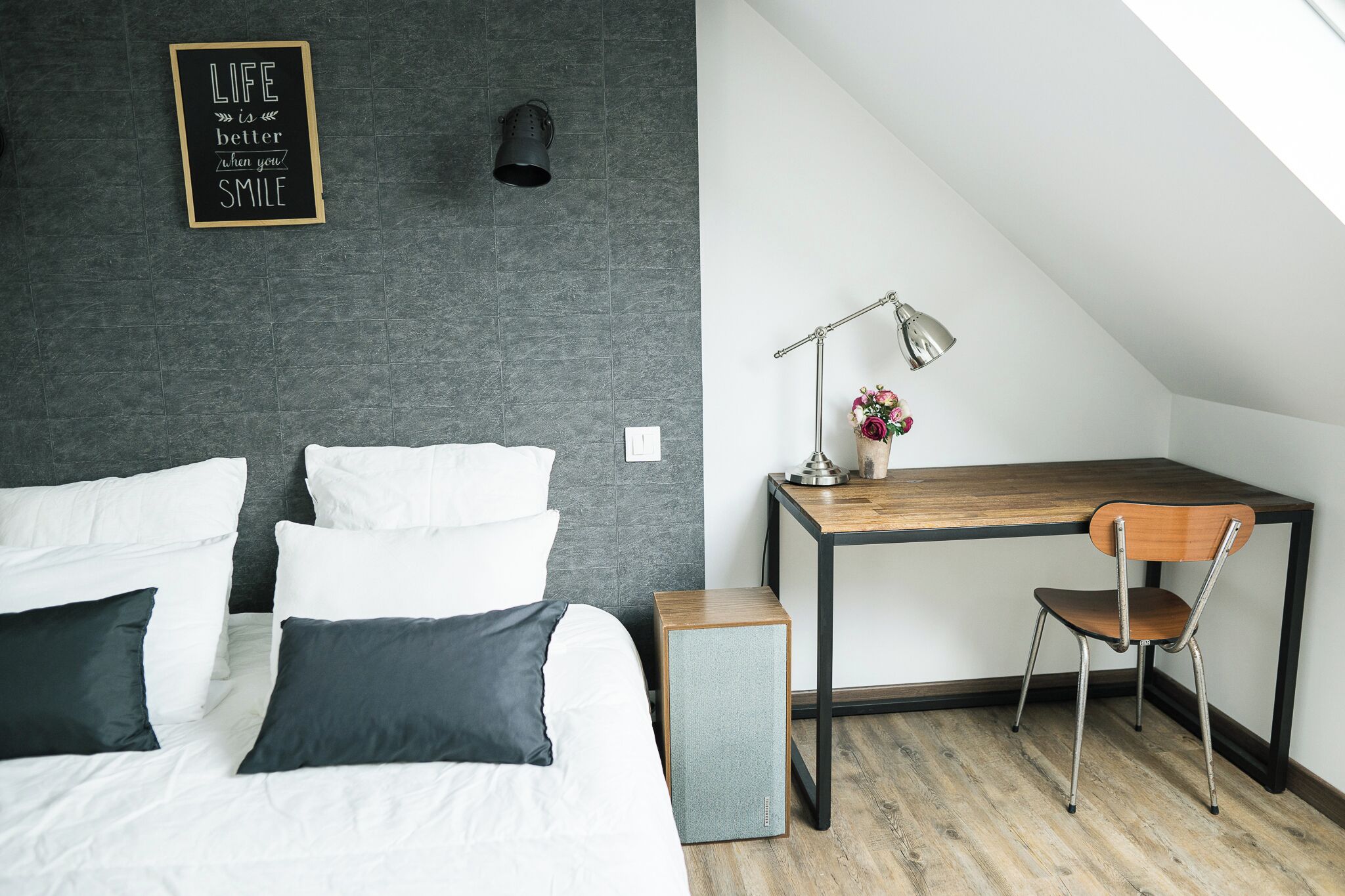 A bedroom with a bed, a desk, and a chair. There is a wall-mounted lamp and a framed quote on the wall.