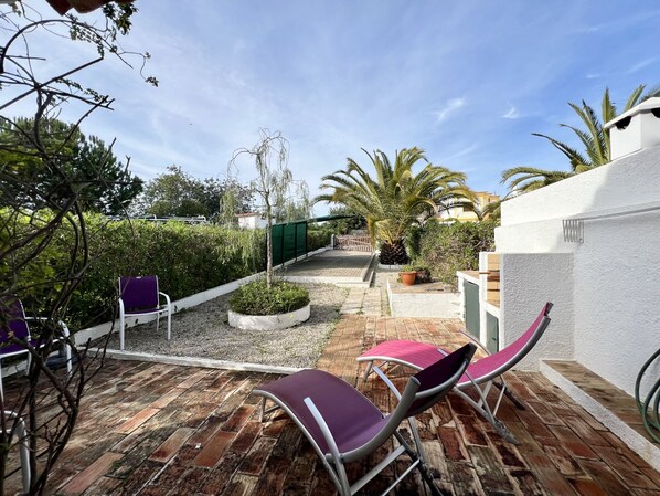 A patio area with a brick floor, two purple lounge chairs, a small garden with a tree and a potted plant, and a white wall with a window.