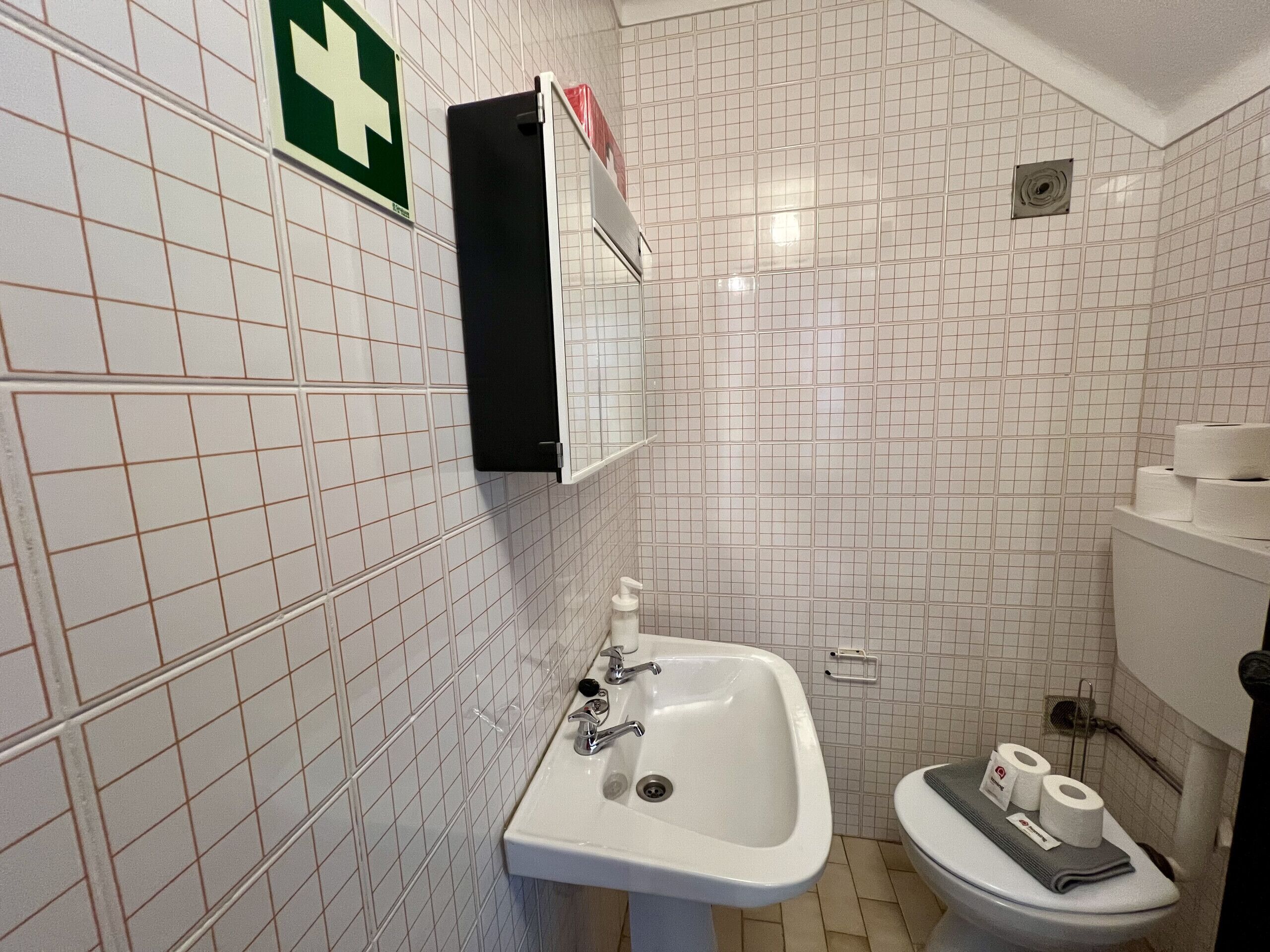 A small bathroom with white tiled walls, a sink, a toilet, and a hand sanitizer dispenser.