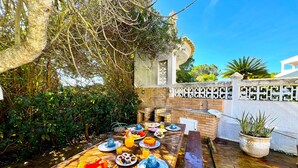 An outdoor dining area with a brick barbecue, surrounded by lush greenery and a clear blue sky. - Vilamoura Brightness Villa by Homing (Loulé)