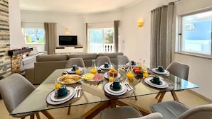 A modern living room with a glass dining table set for breakfast, a sofa, and a TV in the background. - Vilamoura Brightness Villa by Homing (Loulé)