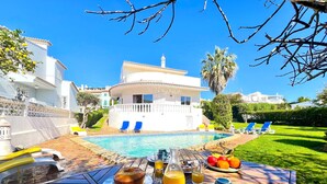 A breakfast table with fruit, juice, and pastries, a pool, and a white villa with blue chairs in the background. - Vilamoura Brightness Villa by Homing (Loulé)