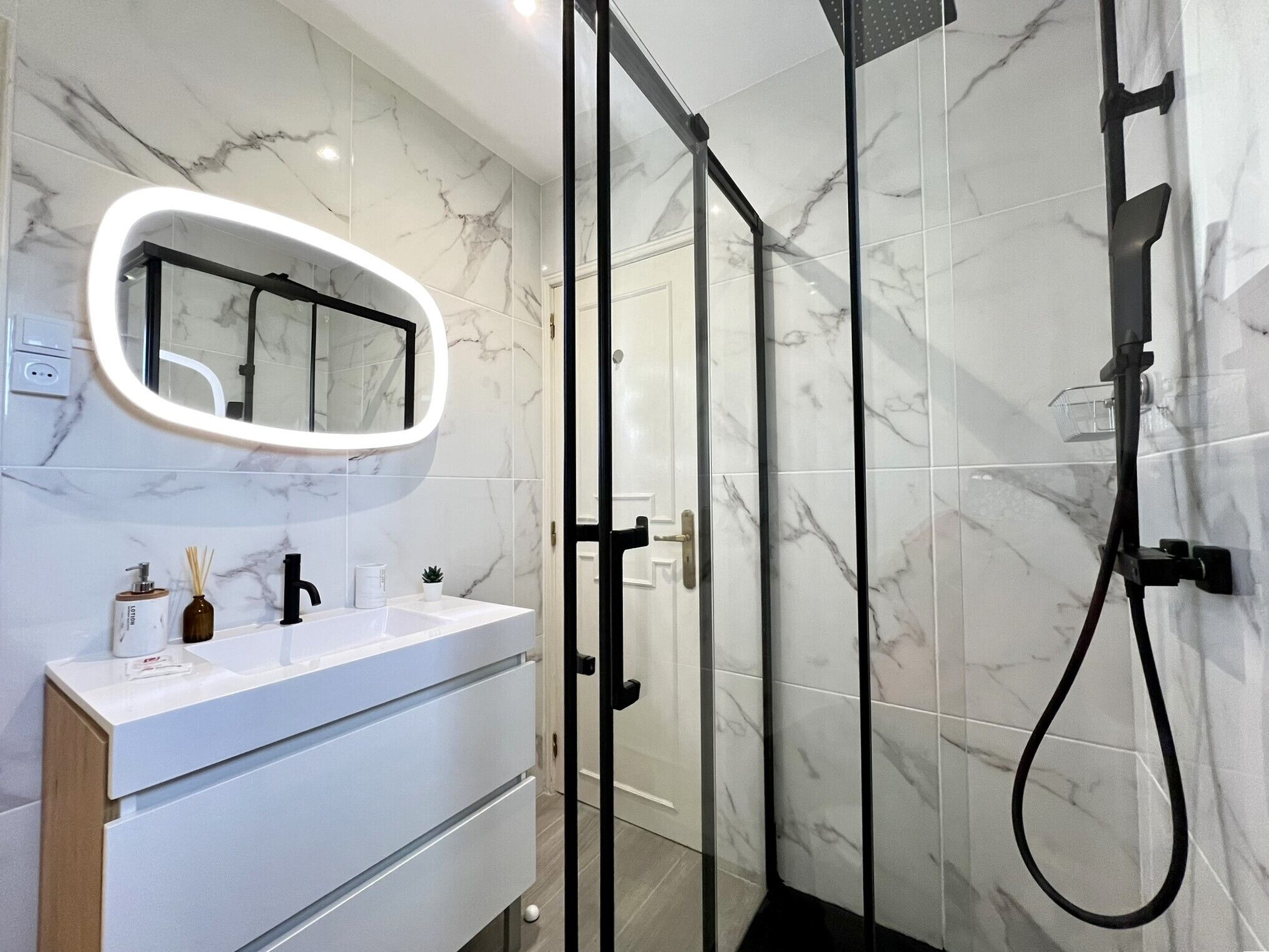 Casa de banho moderna com chuveiro revestido a mármore, área de duche com proteção de vidro e toucador branco com espelho.