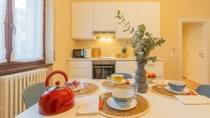Mesa de comedor dispuesta con platos, tazones, vasos y una tetera roja en una cocina luminosa con armarios blancos y una ventana con cortinas translúcidas.
