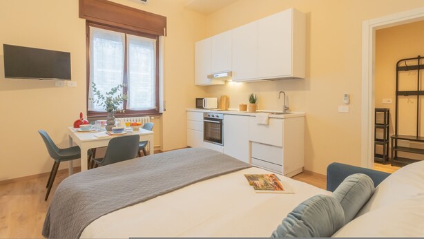 Una habitación compacta con cama, mesita de noche y un pequeño escritorio con silla. Se observa una kitchenette con microondas, estufa y armarios.