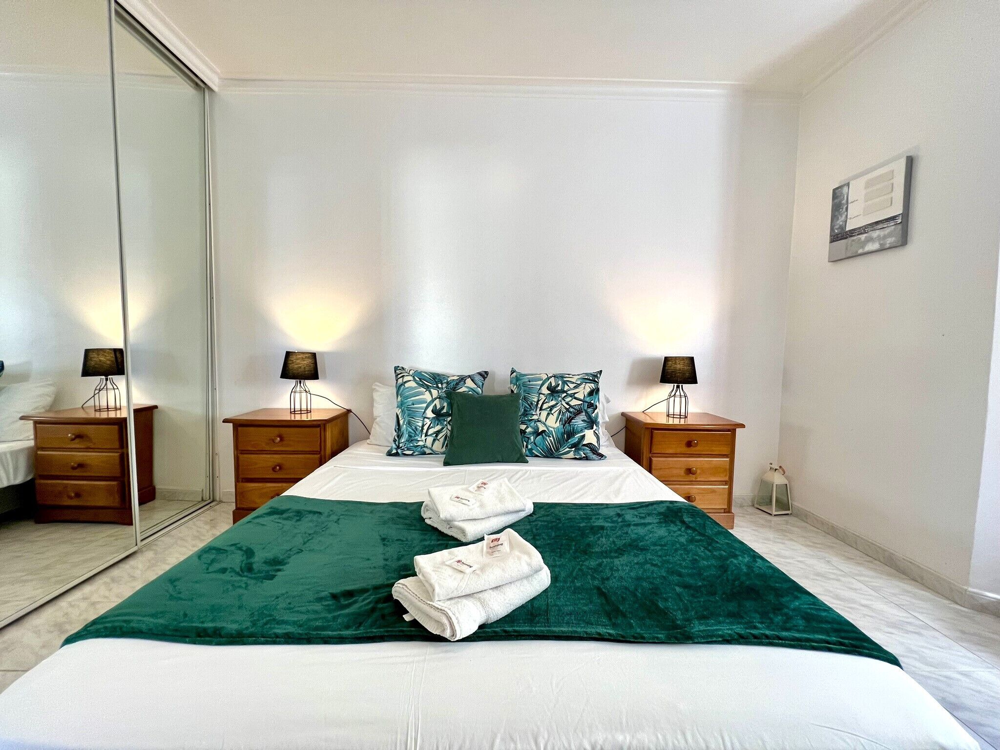 A hotel room with a large bed, two bedside tables with lamps, a mirror, and a green throw blanket.