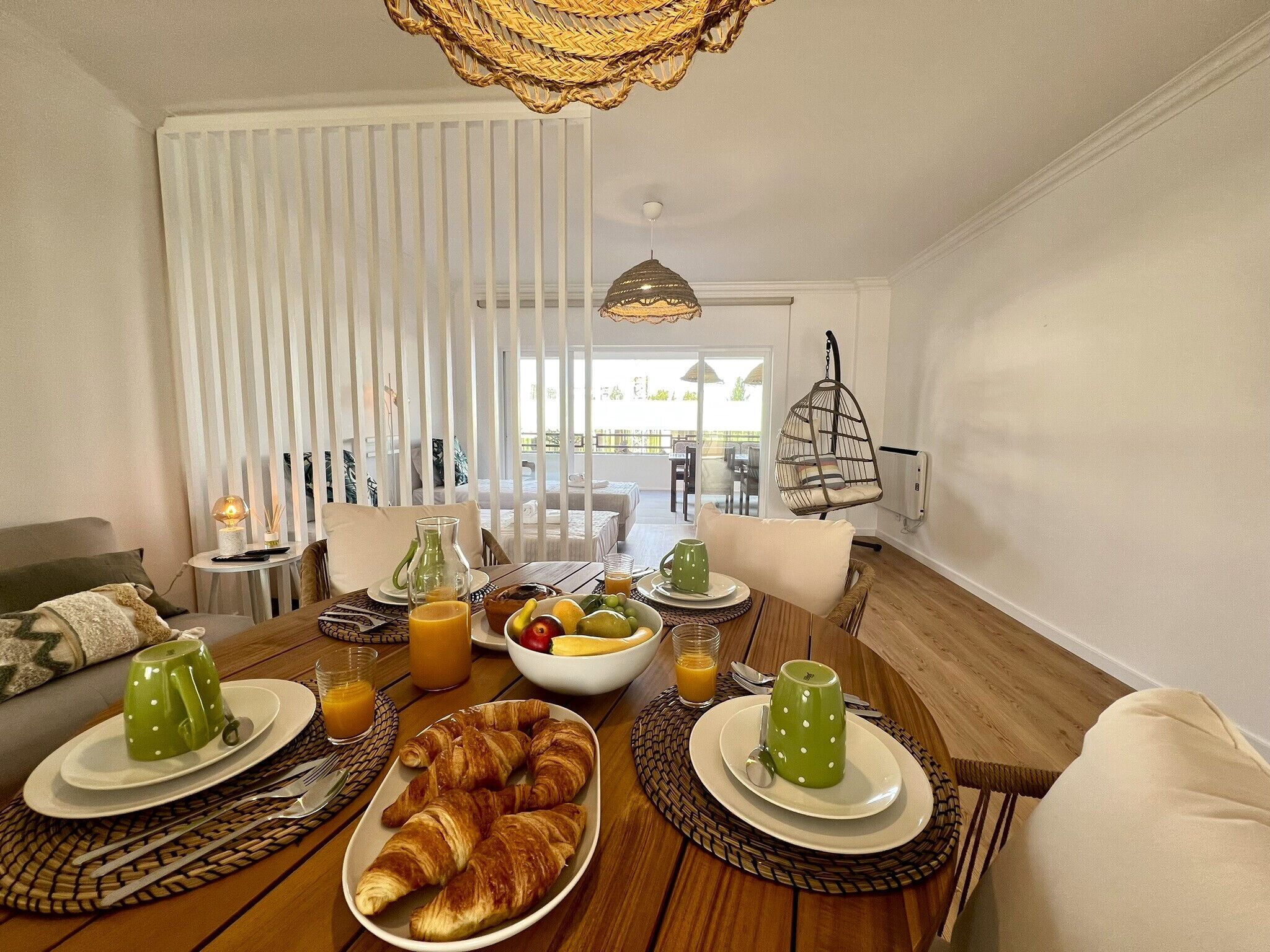 Desayuno con cruasanes, fruta y jugo en una mesa de madera, con vista a un balcón y mesas al aire libre.
