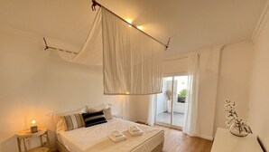 Une chambre avec un grand lit, une table de chevet avec une lampe, un petit tabouret en osier et une vue sur l’extérieur grâce à une porte-fenêtre coulissante.