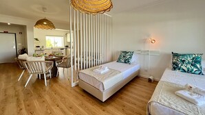 Une chambre d’hôtel moderne avec un plancher en bois, un lit avec une literie blanche, un coin repas avec une table et des chaises, et une cuisine en arrière-plan.