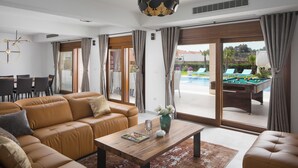 A modern living room with a sofa, coffee table, and a pool table visible through the glass doors. - Malibu Eclipse (Vir)