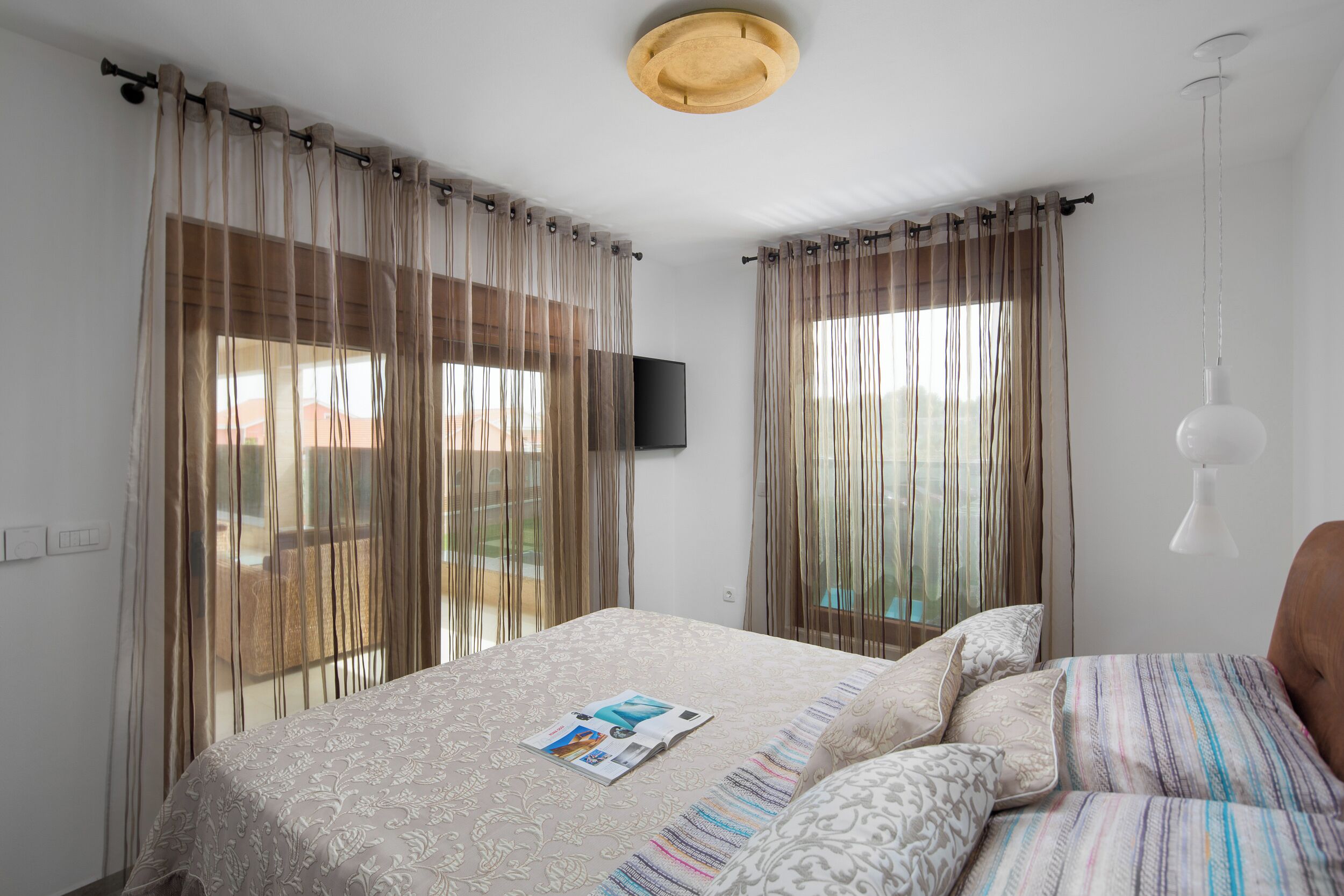 Un dormitorio con cama, cortinas, un televisor montado y una lámpara colgante.
