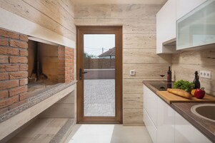 Une cuisine moderne dotée d’un four en briques, d’une porte en bois donnant sur un espace extérieur, et d’un plan de travail sur lequel on trouve une miche de pain, des légumes et une bouteille de vin.